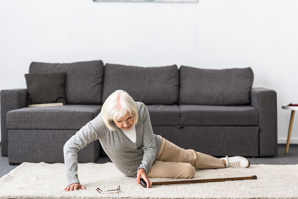 Eldre kvinne med krykke liggjande på golvet framfor ein sofa