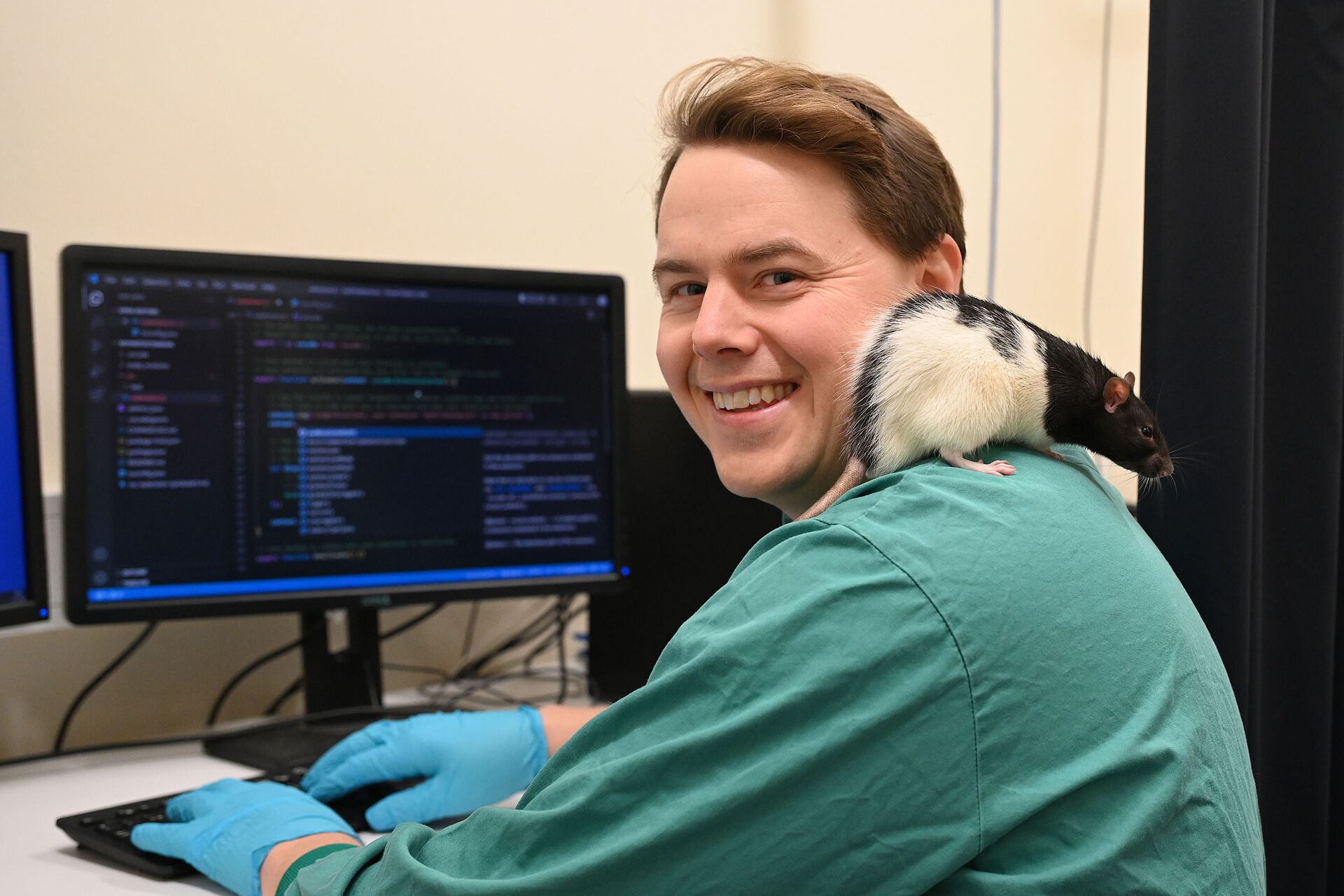 mann i grønne lab-klær sitter ved datamaskin med kode på skjermen og rotte på skulderen
